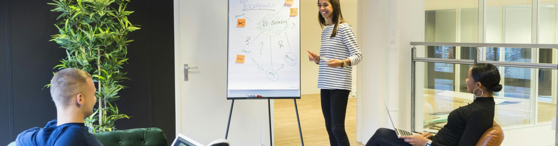 Three adults engaged in a casual office meeting with laptops and a flipchart.
